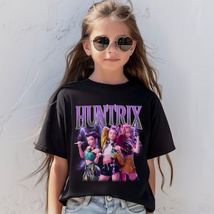 Pu&ograve; includere: T-shirt nera con la scritta "HUNTRIX" in viola e argento, con una grafica di tre donne che tengono dei microfoni. Le donne indossano abiti colorati, con fulmini sullo sfondo.