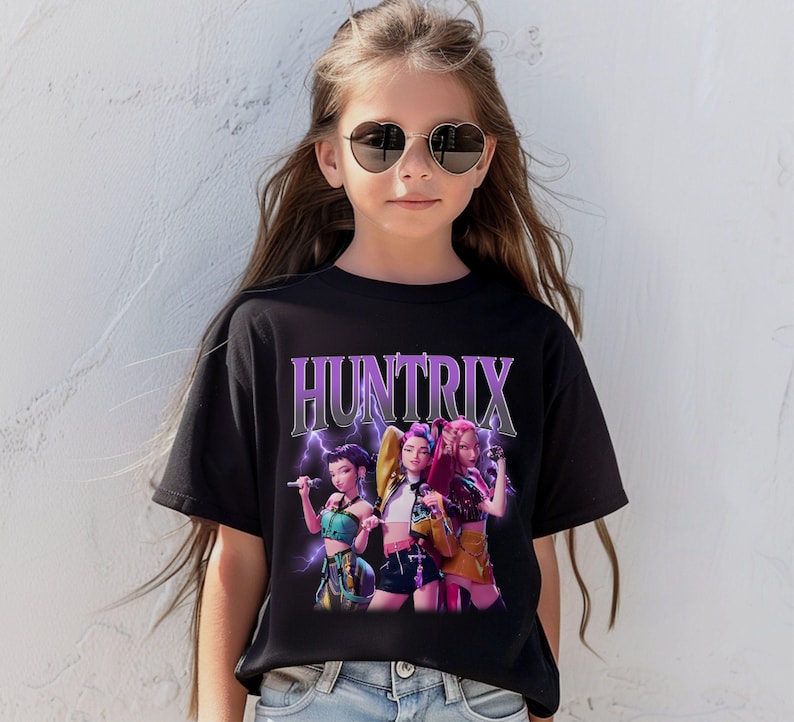 Pu&ograve; includere: T-shirt nera con la scritta "HUNTRIX" in viola e argento, con una grafica di tre donne che tengono dei microfoni. Le donne indossano abiti colorati, con fulmini sullo sfondo.
