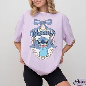 Puede incluir: Una camiseta lavanda con un personaje azul Stitch de Lilo & Stitch. El personaje est&aacute; en un marco con un lazo y la palabra "Ohana!" impresa en la camiseta.