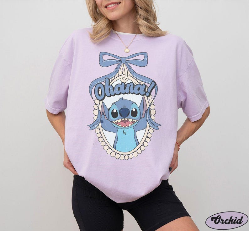 Puede incluir: Una camiseta lavanda con un personaje azul Stitch de Lilo & Stitch. El personaje est&aacute; en un marco con un lazo y la palabra "Ohana!" impresa en la camiseta.