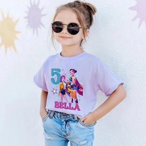 May include: A light purple t-shirt featuring a graphic of animated characters, the number 5, and the name "BELLA" in glittery letters. The shirt is worn by a person wearing sunglasses and blue jeans.