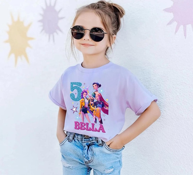 May include: A light purple t-shirt featuring a graphic of animated characters, the number 5, and the name "BELLA" in glittery letters. The shirt is worn by a person wearing sunglasses and blue jeans.