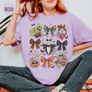 May include: A lilac t-shirt with a Halloween-themed design. The design features illustrations of characters like Jack Skellington and Sally, alongside bow accents and bat motifs. The word "ORCHID" is in the top left corner.