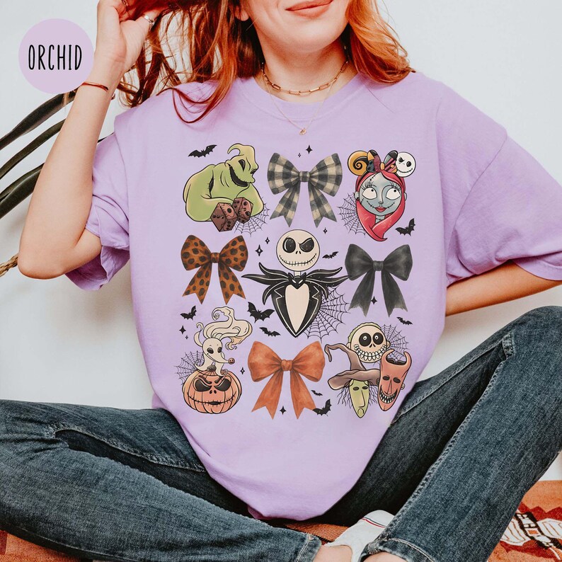 May include: A lilac t-shirt with a Halloween-themed design. The design features illustrations of characters like Jack Skellington and Sally, alongside bow accents and bat motifs. The word "ORCHID" is in the top left corner.