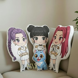 May include: Three cartoon pillows on a beige sofa. The pillows depict anime-style characters with varied hairstyles and outfits. A blue cat pillow is in the centre. The characters have large eyes and are wearing white and gold outfits.