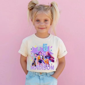 May include: A cream-colored t-shirt featuring a graphic of three animated figures, a large number 5, and the name "MADISON" in a glittery font. The design includes stars and a purple background. The girl in the image is wearing the t-shirt and blue jeans.
