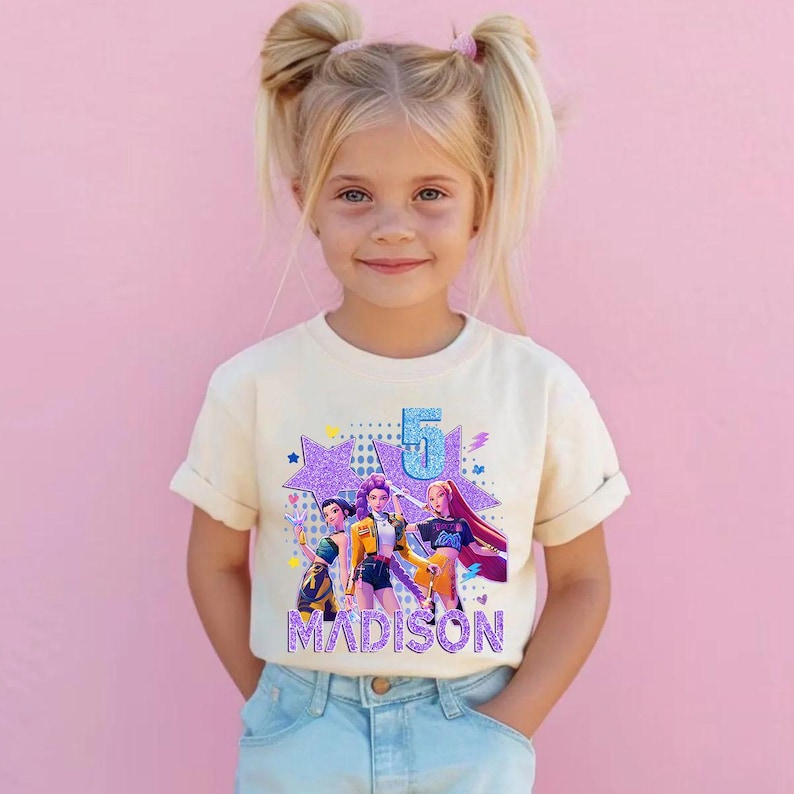 May include: A cream-colored t-shirt featuring a graphic of three animated figures, a large number 5, and the name "MADISON" in a glittery font. The design includes stars and a purple background. The girl in the image is wearing the t-shirt and blue jeans.
