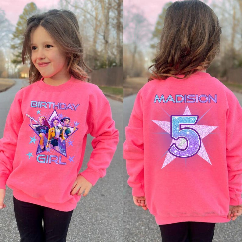 May include: A bright pink sweatshirt with long sleeves. The front features the text "BIRTHDAY GIRL" and a graphic of characters inside a star. The back displays the name "MADISION" and the number "5" inside a star.