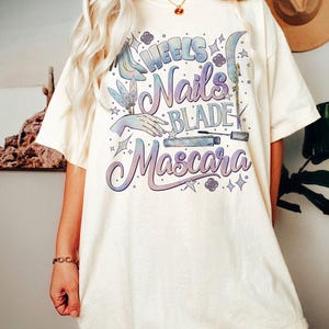 May include: Cream-colored t-shirt with the words "Heels, Nails, Blade, Mascara" in a gradient purple and blue design. The graphic includes illustrations of a high heel, hand, and mascara.