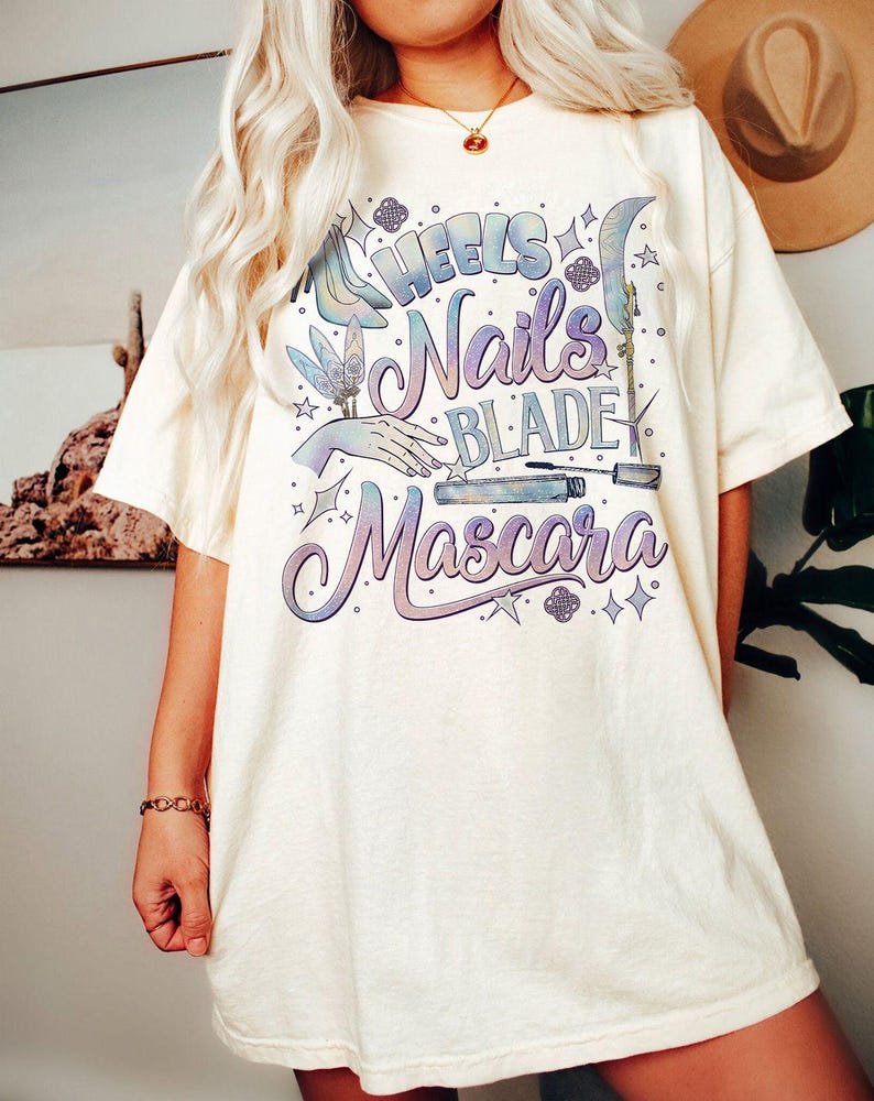 May include: Cream-colored t-shirt with the words "Heels, Nails, Blade, Mascara" in a gradient purple and blue design. The graphic includes illustrations of a high heel, hand, and mascara.