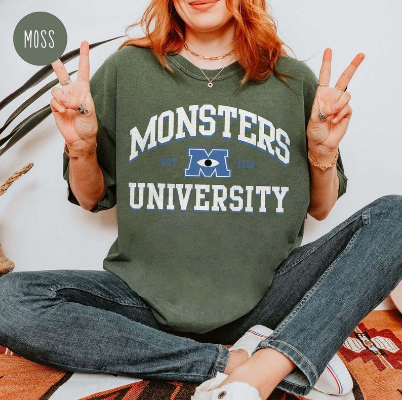 May include: Moss green sweatshirt with the text "MONSTERS UNIVERSITY" in white block letters. The design includes a blue and white eye graphic. The sweatshirt is a casual style, suitable for everyday wear.