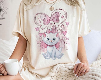 May include: A white t-shirt with a pink and white illustration of a cat wearing a pink bow and standing in front of a pink castle. The illustration is surrounded by pink stars and hearts.