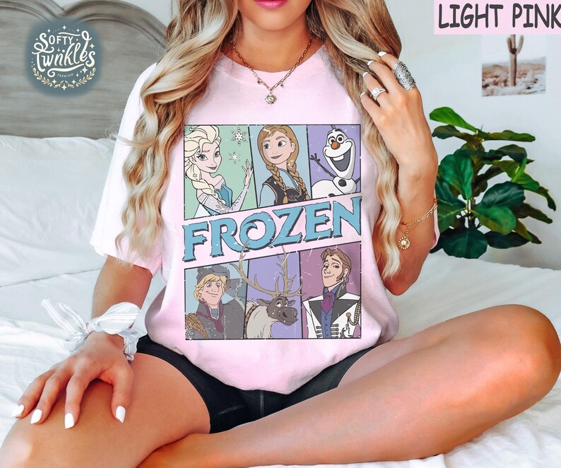 May include: A light pink t-shirt with a vintage-style graphic of characters from the movie Frozen. The graphic features Elsa, Anna, Kristoff, Olaf, and Sven. The text "FROZEN" is printed in blue letters.