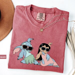 May include: Crimson red t-shirt with a graphic print of Stitch and Lilo from the Disney movie Lilo & Stitch. Stitch is wearing a green grass skirt and sunglasses. Lilo is wearing a pink dress and sunglasses. The shirt has a tag that says "Comfort Colors" and "S-P-CH".