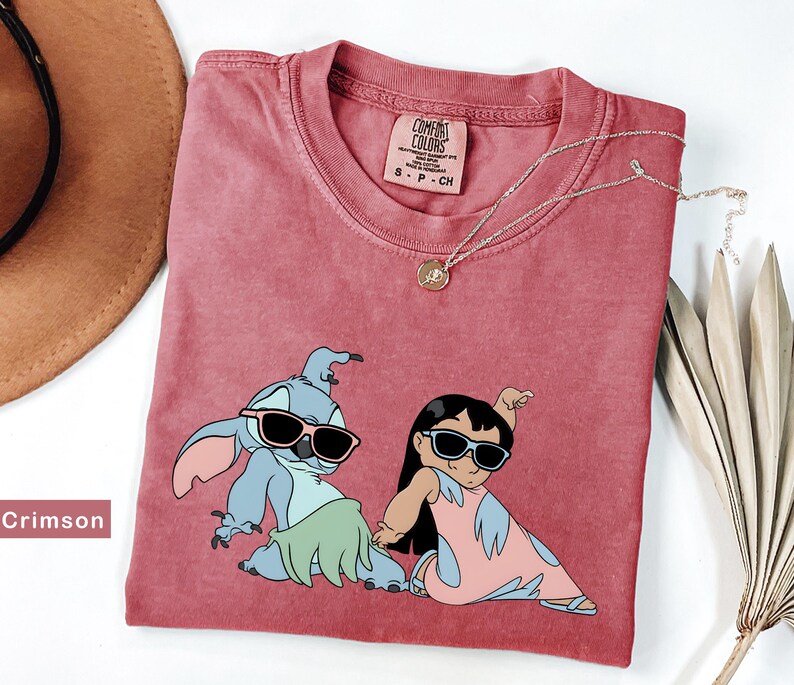 May include: Crimson red t-shirt with a graphic print of Stitch and Lilo from the Disney movie Lilo & Stitch. Stitch is wearing a green grass skirt and sunglasses. Lilo is wearing a pink dress and sunglasses. The shirt has a tag that says "Comfort Colors" and "S-P-CH".