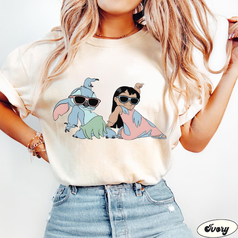 May include: A white t-shirt with a cartoon image of Stitch and Lilo from the Disney movie Lilo & Stitch. Stitch is wearing sunglasses and a green lei, while Lilo is wearing a pink dress and sunglasses. The shirt is a light ivory color.