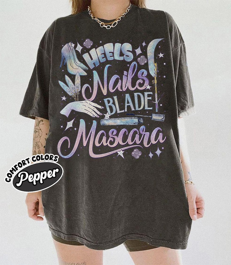 May include: Dark gray oversized t-shirt with the words "Heels, Nails, Blade, Mascara" in a holographic design. The shirt features illustrations of a high heel, hand, and mascara wand. The brand name "Comfort Colors Pepper" is printed on the shirt.