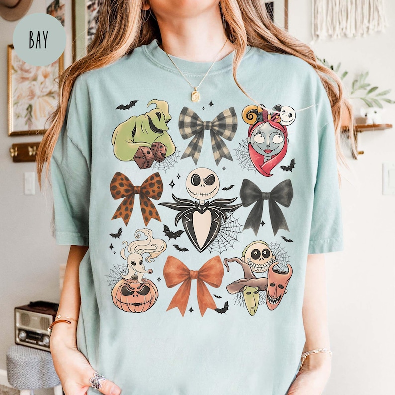 May include: A pale blue t-shirt with a graphic design featuring characters and bow designs. The design includes Jack Skellington, Sally, and Oogie Boogie, along with various bow patterns and spiderwebs.