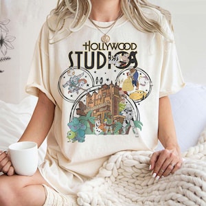 Può includere: T-shirt color crema con grafica di Hollywood Studios e personaggi dei cartoni animati. Il design include il testo "Hollywood Studios" e immagini di personaggi di vari film d'animazione. La maglietta ha maniche corte e una vestibilità ampia.