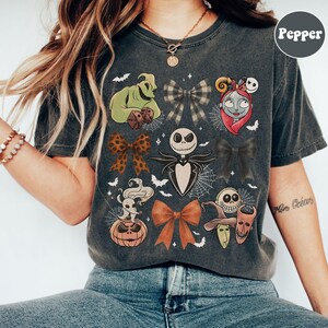 May include: Dark grey t-shirt featuring various character designs and bows from a popular animated film. The shirt includes images of Jack Skellington, Sally, Oogie Boogie, and other characters. The word "Pepper" is printed on the upper right.