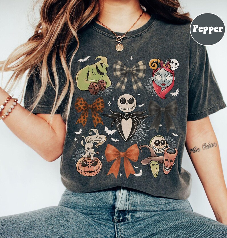 May include: Dark grey t-shirt featuring various character designs and bows from a popular animated film. The shirt includes images of Jack Skellington, Sally, Oogie Boogie, and other characters. The word "Pepper" is printed on the upper right.