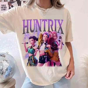 Pu&ograve; includere: T-shirt color crema con la scritta "HUNTRIX" in un font sfumato viola sopra una grafica di tre figure animate che tengono in mano dei microfoni. Le figure indossano abiti di scena colorati, con fulmini sullo sfondo.