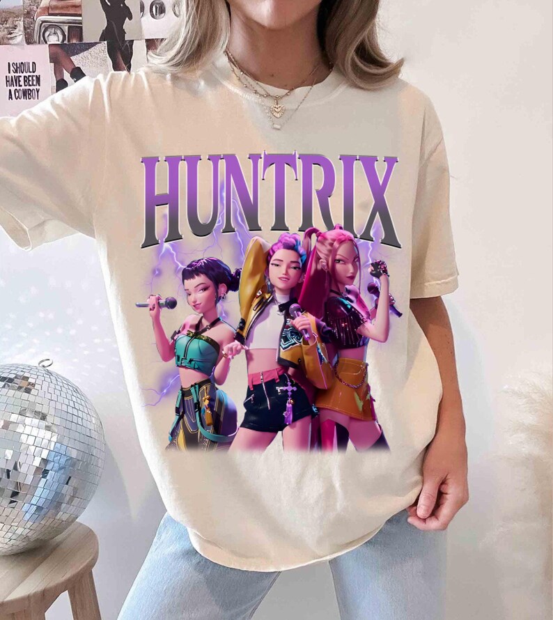 Pu&ograve; includere: T-shirt color crema con la scritta "HUNTRIX" in un font sfumato viola sopra una grafica di tre figure animate che tengono in mano dei microfoni. Le figure indossano abiti di scena colorati, con fulmini sullo sfondo.