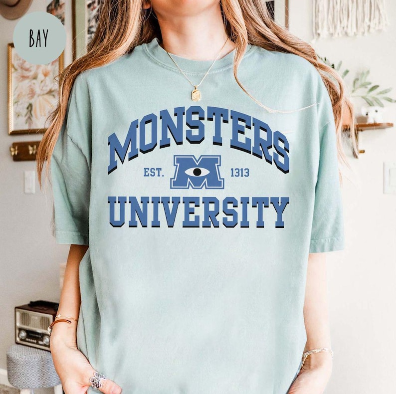May include: A light blue t-shirt with the text "MONSTERS UNIVERSITY" in blue. The design includes an eye graphic and the text "EST. 1313". The shirt has short sleeves and a crew neck.
