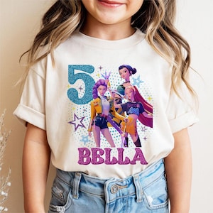 May include: A cream-colored t-shirt featuring a graphic design with the number "5" in blue glitter, the name "BELLA" in pink glitter, and illustrations of three animated female characters. The shirt is worn by a person.