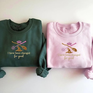 以下が含まれることがあります： 刺繍デザインが施された、ダークグリーンとピンクの2つのクルーネックのスウェットシャツ。それぞれに魔女の帽子、ほうき、王冠、そして「I have been changed for good」の文字が金色で刺繍されています。スウェットシャツはカジュアルウェアです。