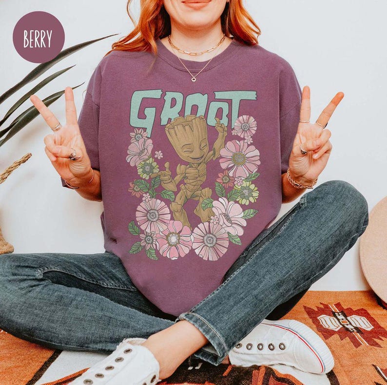 May include: A purple t-shirt featuring a graphic of Groot from Guardians of the Galaxy. The graphic shows Groot standing in a field of flowers. The text "Groot" is printed in green above the graphic.