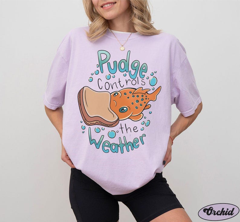 May include: A lavender t-shirt featuring a cartoon illustration of a fish with a slice of bread on its head. The text on the shirt reads "Pudge controls the weather".