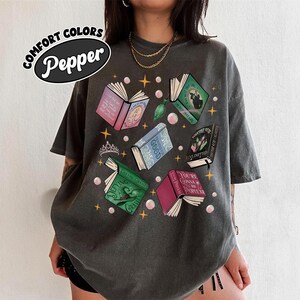 May include: Dark grey t-shirt with a graphic of open books in various colors, with titles like "Defying Gravity" and "You're Gonna Be Popular." The shirt also features the text "COMFORT COLORS Pepper."