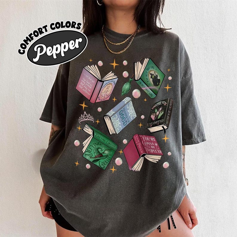 May include: Dark grey t-shirt with a graphic of open books in various colors, with titles like "Defying Gravity" and "You're Gonna Be Popular." The shirt also features the text "COMFORT COLORS Pepper."