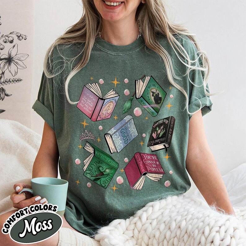 May include: Moss green Comfort Colors t-shirt featuring a graphic of open books with various titles and illustrations. The design includes pink, blue, and green books, along with stars and bubbles. The shirt has rolled-up sleeves.