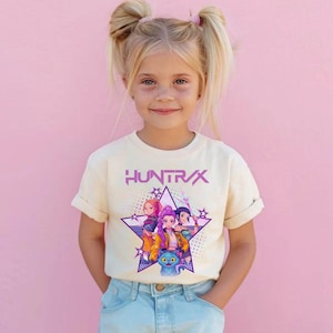 Pode incluir: Uma camiseta creme com a palavra "HUNTRX" em roxo, acima de um gr&aacute;fico de quatro personagens de desenho animado dentro de uma estrela roxa. Os personagens s&atilde;o de v&aacute;rias cores. A camiseta &eacute; usada por uma menina.