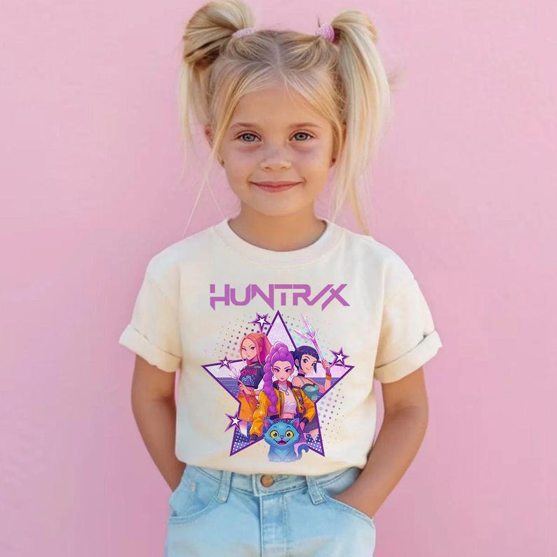 Pode incluir: Uma camiseta creme com a palavra "HUNTRX" em roxo, acima de um gr&aacute;fico de quatro personagens de desenho animado dentro de uma estrela roxa. Os personagens s&atilde;o de v&aacute;rias cores. A camiseta &eacute; usada por uma menina.