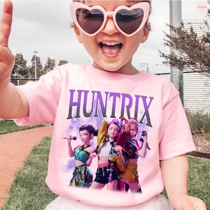 Pu&ograve; includere: T-shirt rosa chiaro con la scritta "HUNTRIX" in viola e una grafica di tre personaggi animati con microfoni. I personaggi indossano abiti colorati. Lo sfondo presenta dei fulmini.