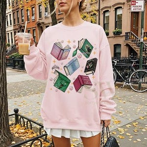 May include: A light pink sweatshirt featuring a design of open books in various colors, including pink, green, and blue. The books are surrounded by stars and bubbles. The sweatshirt is paired with a white skirt.