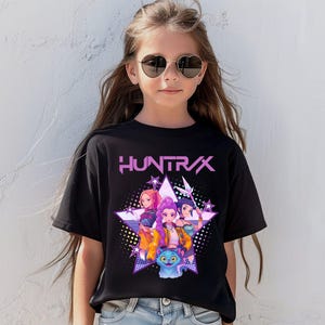 Pode incluir: T-shirt preto com a palavra "HUNTRAX" em rosa e um gr&aacute;fico de tr&ecirc;s meninas animadas e um gato azul dentro de uma estrela. As meninas usam roupas coloridas. A t-shirt &eacute; usada por uma pessoa que usa &oacute;culos de sol em forma de cora&ccedil;&atilde;o.