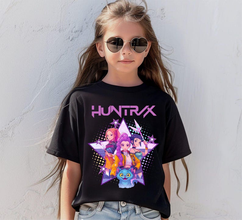 Pode incluir: T-shirt preto com a palavra "HUNTRAX" em rosa e um gr&aacute;fico de tr&ecirc;s meninas animadas e um gato azul dentro de uma estrela. As meninas usam roupas coloridas. A t-shirt &eacute; usada por uma pessoa que usa &oacute;culos de sol em forma de cora&ccedil;&atilde;o.