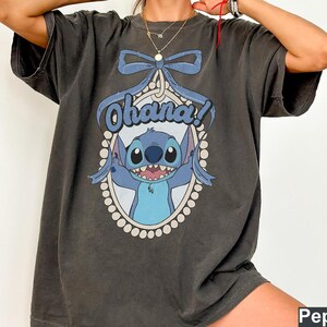 Puede incluir: Una camiseta gris oscuro con un personaje azul Stitch de Lilo & Stitch. El personaje est&aacute; en un marco con un borde de perlas y el texto "Ohana!" encima.
