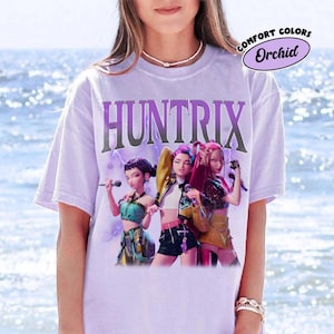 Pu&ograve; includere: Una maglietta lilla chiaro con la scritta "HUNTRIX" in un carattere stilizzato sopra una grafica di tre donne. Le donne tengono in mano dei microfoni e indossano abiti colorati. La maglietta ha un'etichetta "Comfort Colors" con la parola "Orchid".