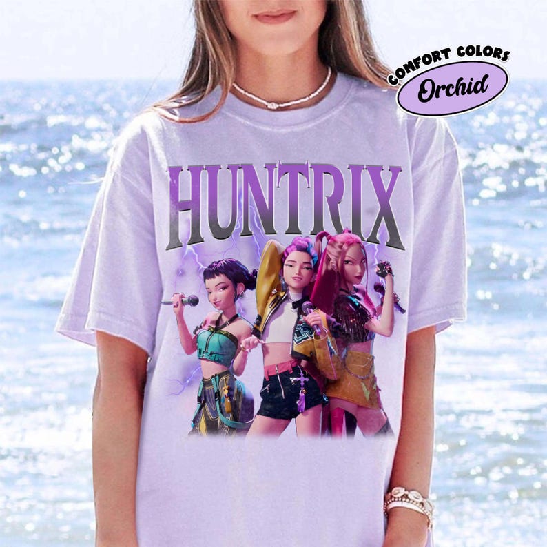 Pu&ograve; includere: Una maglietta lilla chiaro con la scritta "HUNTRIX" in un carattere stilizzato sopra una grafica di tre donne. Le donne tengono in mano dei microfoni e indossano abiti colorati. La maglietta ha un'etichetta "Comfort Colors" con la parola "Orchid".