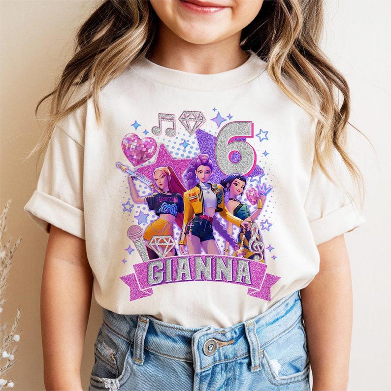 May include: A cream-colored t-shirt featuring a colorful graphic of three animated figures, a microphone, musical notes, and the number 6. The name "GIANNA" is displayed on a banner below the figures. The design includes glitter accents.