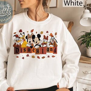 May include: White sweatshirt with a graphic of Mickey Mouse, Minnie Mouse, Donald Duck, Goofy, and Pluto in front of a sign that says "Disneyland". The characters are wearing fall-themed outfits and there are autumn leaves scattered around them.