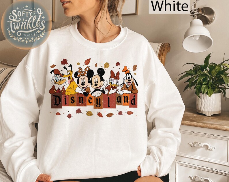 May include: White sweatshirt with a graphic of Mickey Mouse, Minnie Mouse, Donald Duck, Goofy, and Pluto in front of a sign that says "Disneyland". The characters are wearing fall-themed outfits and there are autumn leaves scattered around them.