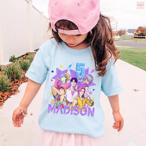 May include: A light blue t-shirt featuring a colorful graphic of anime-style characters, the number 5, and the name "MADISON" in purple. The shirt is worn by a young child wearing a pink baseball cap.