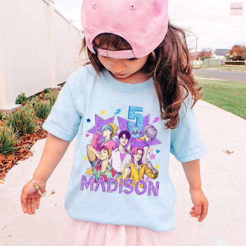 May include: A light blue t-shirt featuring a colorful graphic of anime-style characters, the number 5, and the name "MADISON" in purple. The shirt is worn by a young child wearing a pink baseball cap.