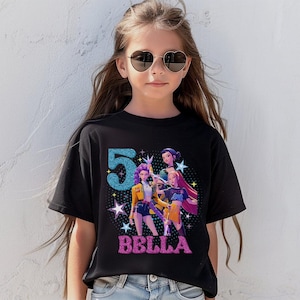 May include: Black t-shirt featuring a colorful graphic design with the number "5" in blue glitter and the name "BELLA" in pink glitter. The graphic includes illustrations of animated characters. The shirt is worn by a young person.
